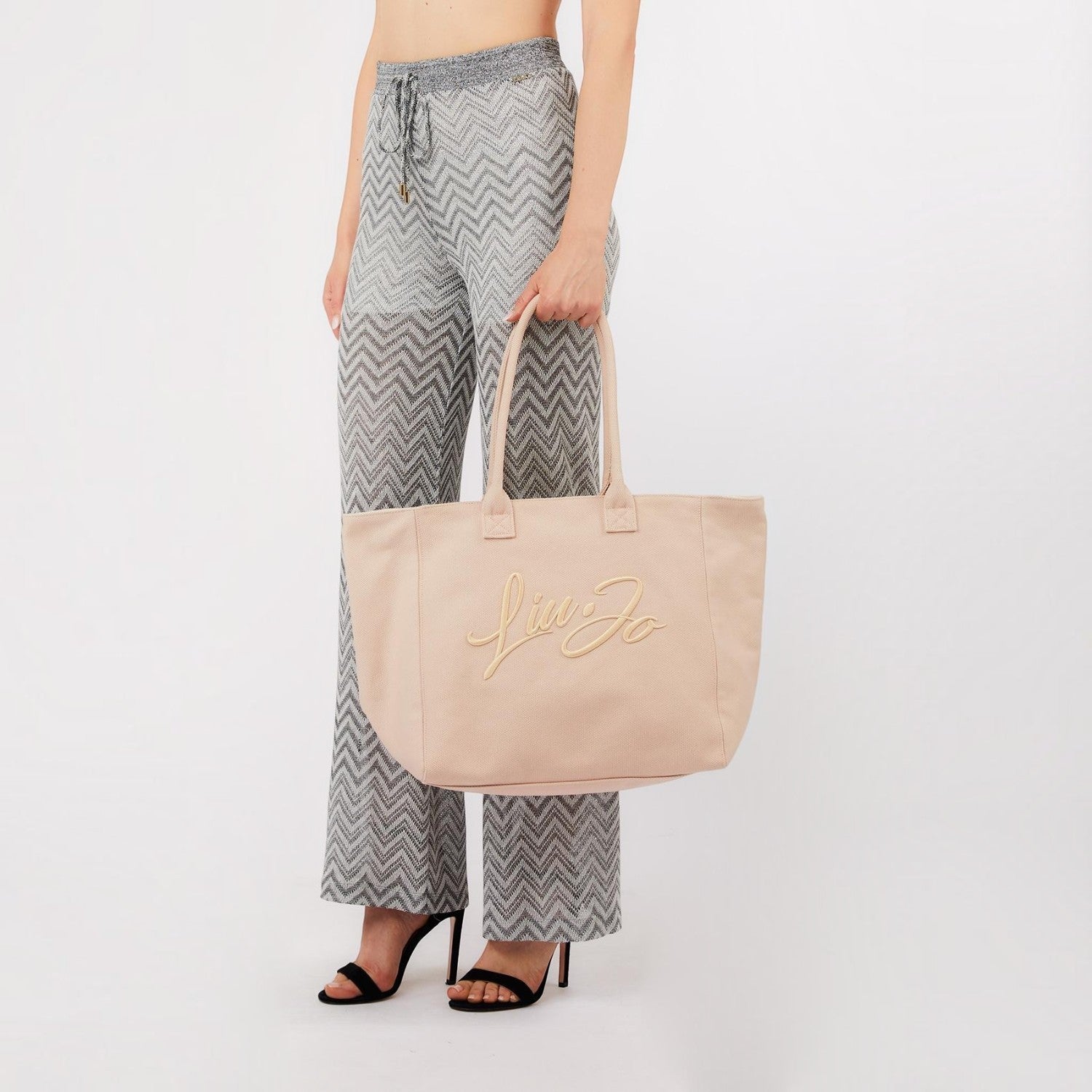 Lieblingstasche Tote L in beige canvas with embroidered logo, held by a woman wearing chevron patterned trousers.