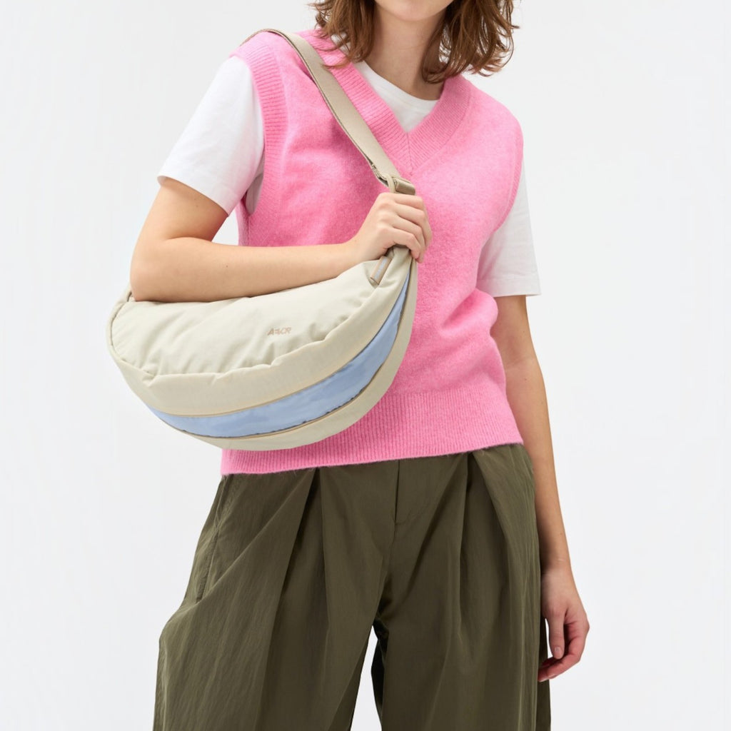 Lieblingstasche beige crescent sling bag with a light blue accent stripe worn by a woman in a pink sweater vest