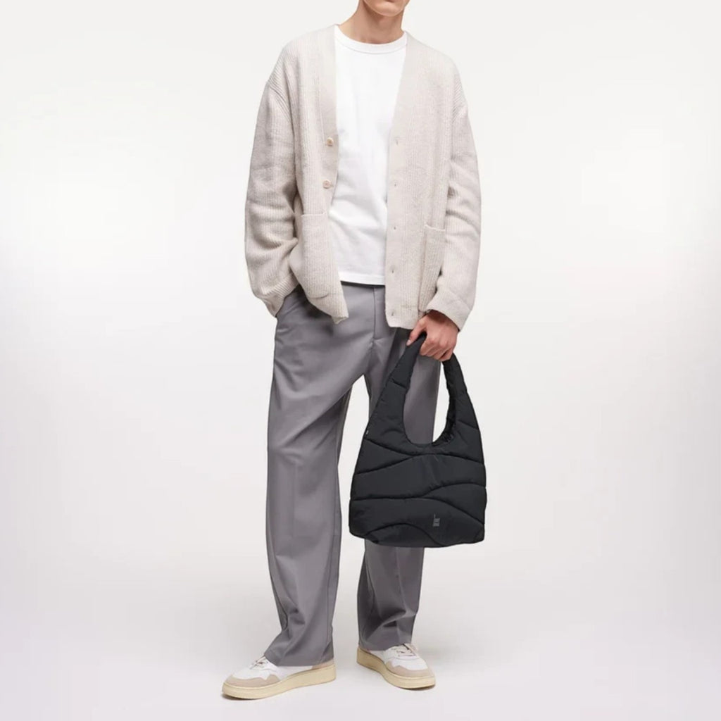 A male model holding a black Lieblingstasche wavy puffer shoulder bag while wearing a beige cardigan and grey trousers.