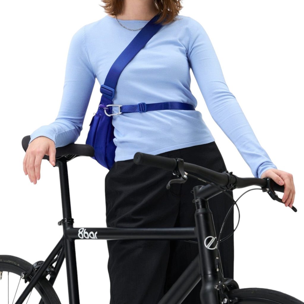 Woman wearing a bright blue Lieblingstasche sling bag across her chest while leaning on a black bicycle