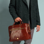 Lieblingstasche brown leather briefcase with gold hardware, held by a man in a grey turtleneck and charcoal coat.