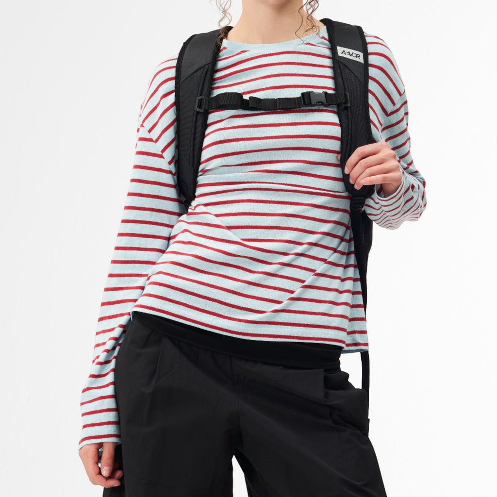 A smiling woman wearing a black Lieblingstasche Day Pack FLX backpack over a light blue and red striped long-sleeve top
