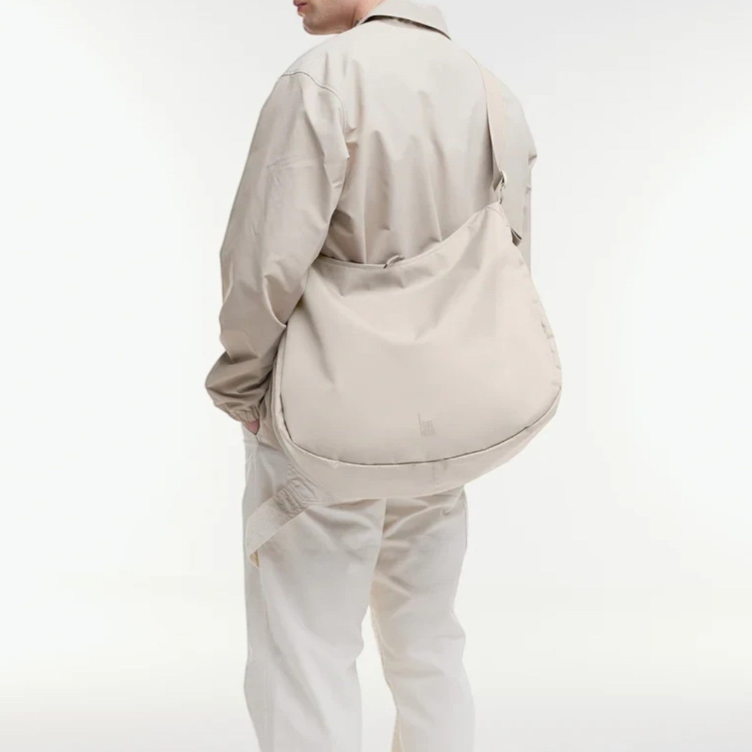 Lieblingstasche GOT BAG beige curved crossbody bag worn by a man in a matching light outfit against a white background