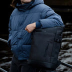 Man wearing a blue puffer jacket and black beanie posing with a Lieblingstasche Helsinki black minimalist backpack