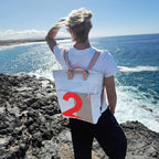 Lieblingstasche Kaje white canvas backpack with tan accents and a large orange number 2 graphic, worn by a woman overlooking the ocean.