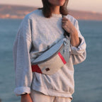 Lieblingstasche Knoten belt bag made from recycled white sailcloth with red accents worn by a woman at the coast.