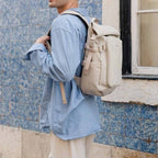 Lieblingstasche Lisbon small beige rolltop backpack worn by a man in a blue striped shirt against a blue tiled wall.