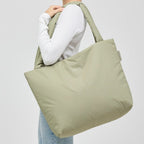 A woman wearing a sage green Lieblingstasche Norva 2in1 shopper bag over her shoulder against a light grey background.