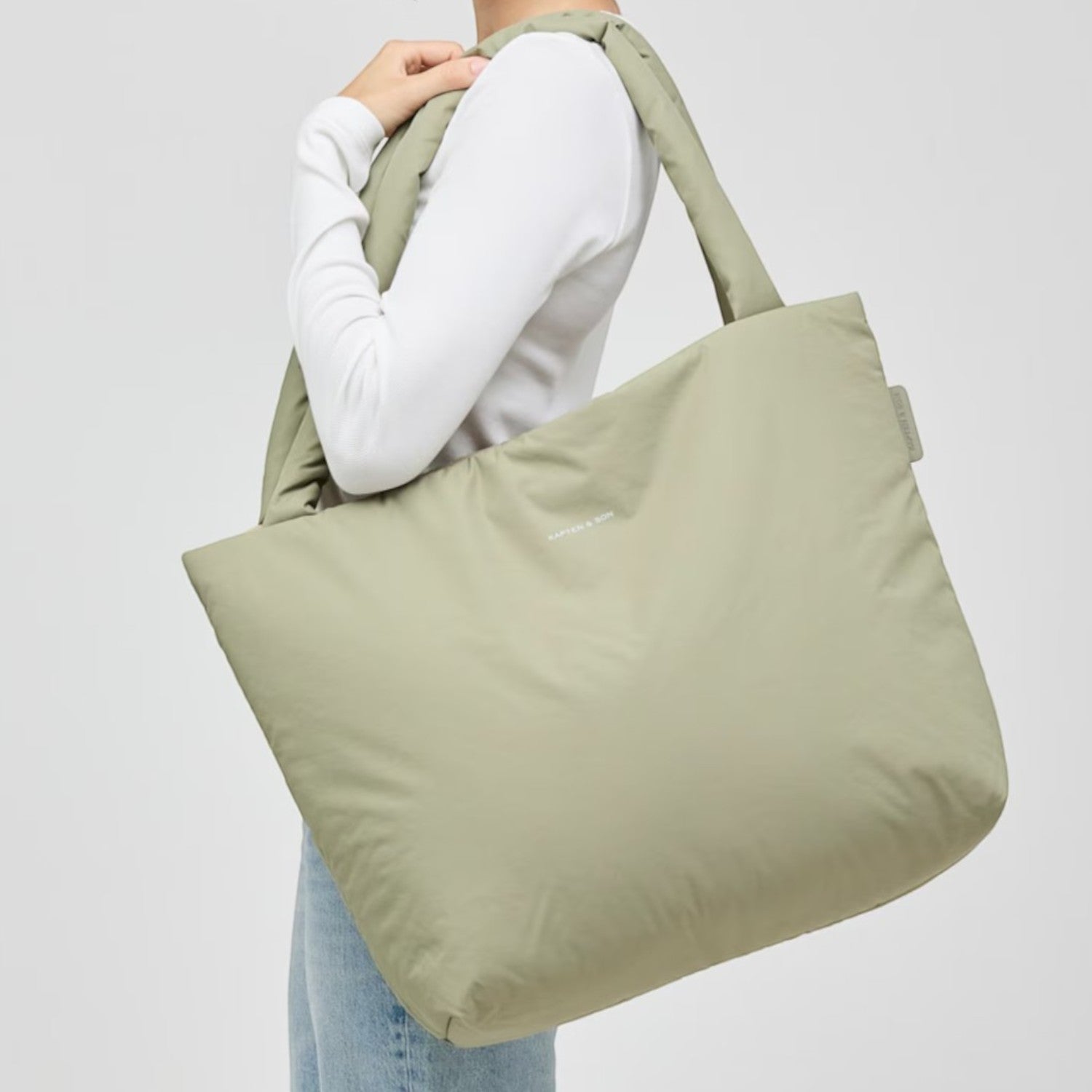 A woman wearing a sage green Lieblingstasche Norva 2in1 shopper bag over her shoulder against a light grey background.