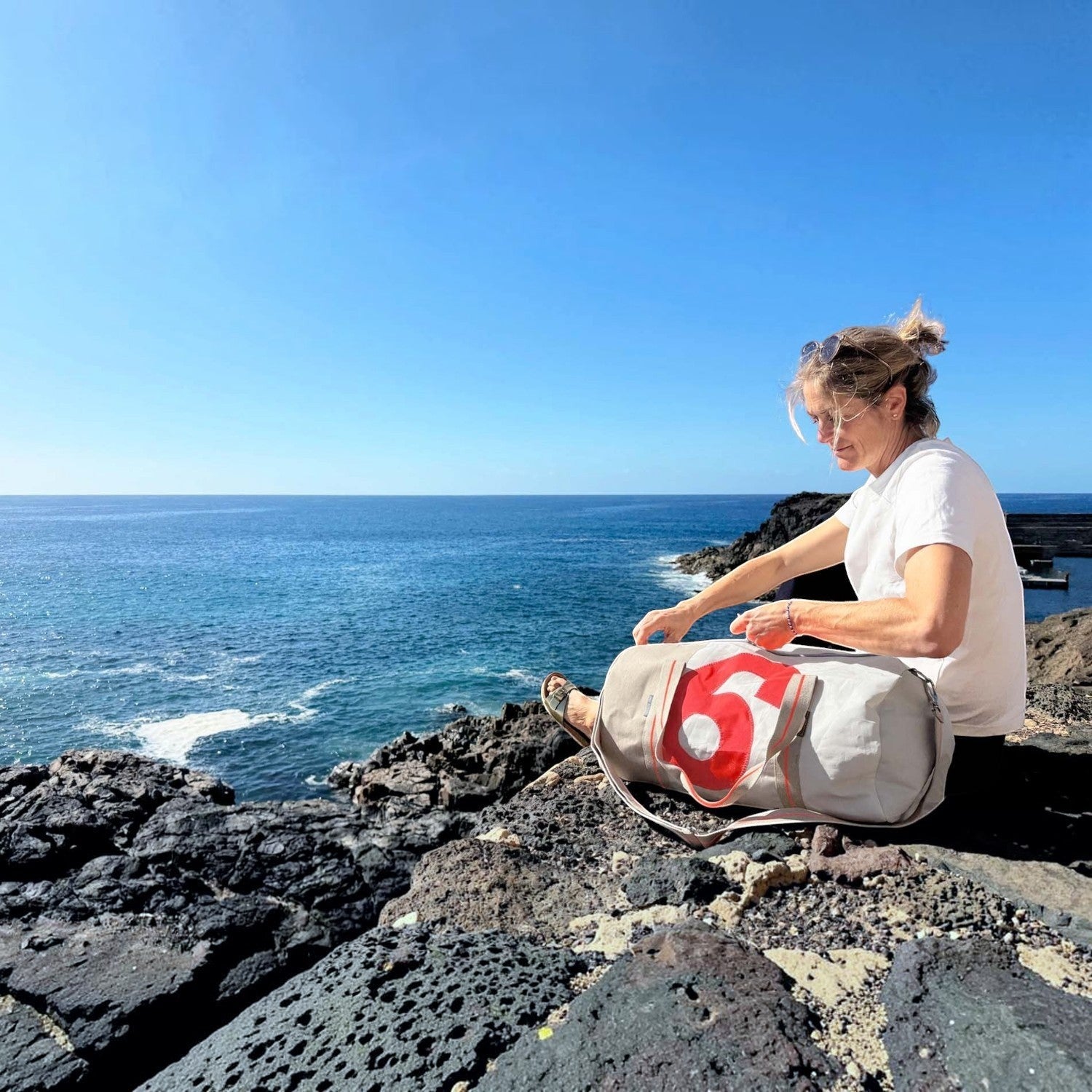 Woman sitting on a rocky cliff by the sea with a Lieblingstasche Pirat grey duffel bag featuring a red number six