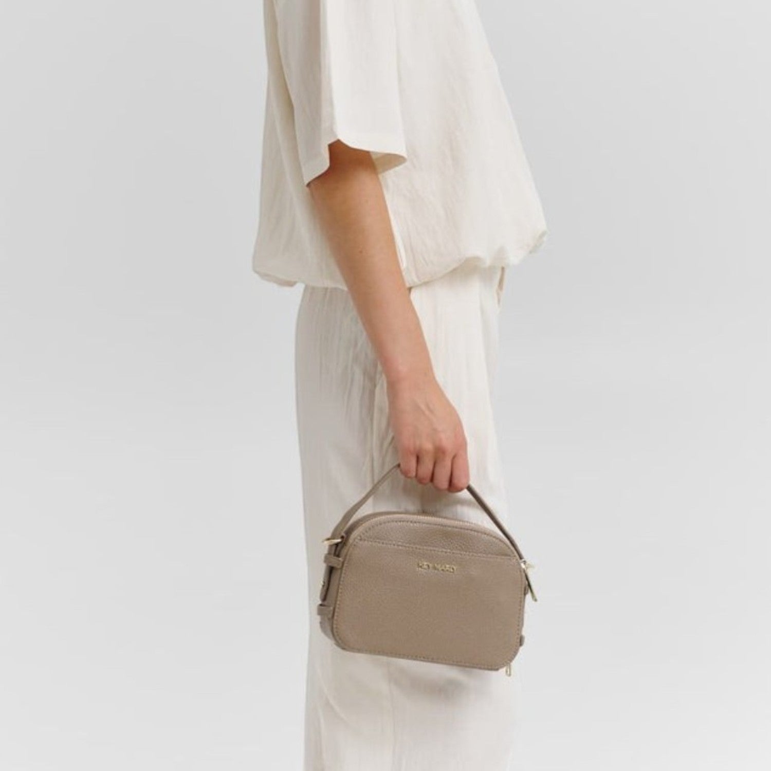 A woman wearing a cream-colored outfit holding a taupe Lieblingstasche Style Mate crossbody bag.