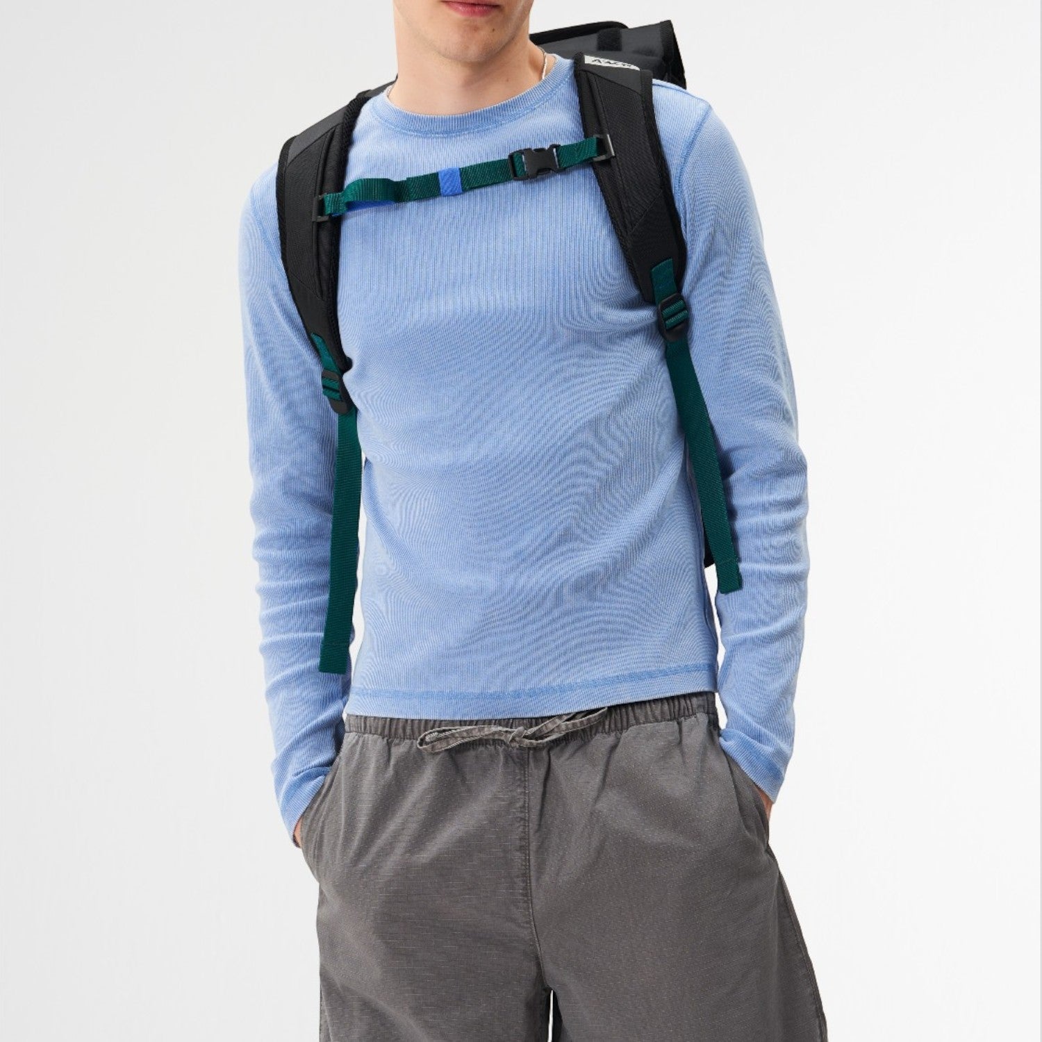 A man wearing a black Lieblingstasche Trip Pack backpack with green and blue straps over a light blue long-sleeve shirt