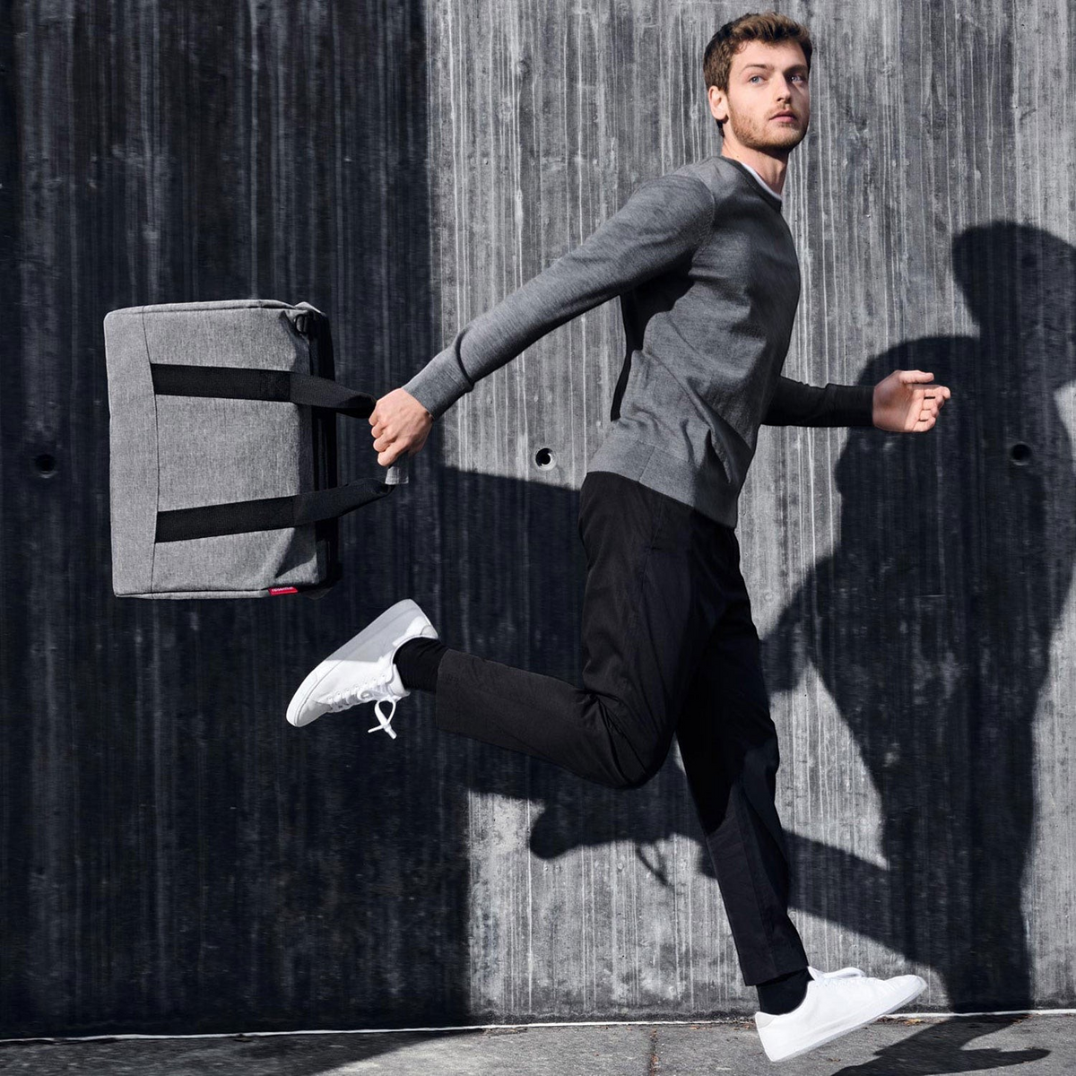 Lieblingstasche Allrounder L grey travel bag held by a man in a grey sweater running against a concrete wall